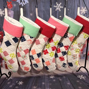 Puede incluir: Un conjunto de seis calcetines navideños acolchados con coloridos diseños de patchwork. Cada calcetín tiene un puño de diferente color, incluyendo rosa, verde y rojo con lunares blancos. Los calcetines se exhiben en un soporte de metal negro.