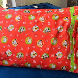 May include: A rectangular pillowcase with a festive Christmas theme. The red fabric is patterned with Mickey Mouse in Santa hats and ornaments. A green stripe with Mickey Mouse designs accents the side. The pillowcase is on a blue surface.