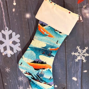 Op de afbeelding: Een gewatteerde kerstkous met een levendige print van een berglandschap met skiërs, besneeuwde bergtoppen en feloranje zonsondergangwolken. De kous heeft een effen crèmekleurige boord aan de bovenkant en een bijpassende stoffen lus om op te hangen.