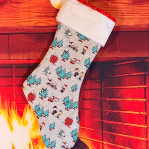May include: A gray Christmas stocking featuring a repeating pattern of Snoopy, Charlie Brown, and other Peanuts characters among teal pine trees. The stocking has a white plush cuff with a red and white polka dot trim and a white pom-pom.