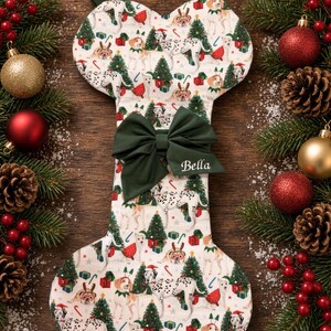 May include: A bone-shaped Christmas stocking featuring a white fabric base with a repeating pattern of dogs, Christmas trees, and gifts. A dark green fabric bow is attached to the center, with the name Bella embroidered in white script.