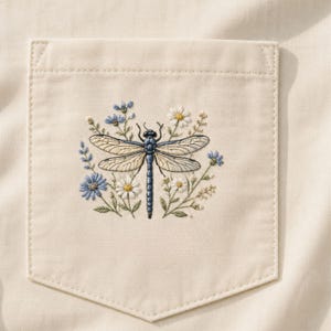 Puede incluir: Bolsillo color crema con un diseño de libélula bordada. La libélula es azul y beige, rodeada de flores azules y blancas y follaje verde. El bolsillo está cosido sobre una tela color crema.