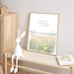 May include: A framed watercolor print with the text "Long Live all the magic we made" above a field of wildflowers. A white rabbit figurine sits on a bench next to the print. A small wooden house and a rainbow-shaped toy are also visible.