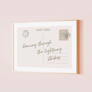 May include: A framed postcard print with a gold frame. The postcard has a light beige background with the words "Dancing through the lightning strikes" in a handwritten font. It includes a stamp and a postmark.