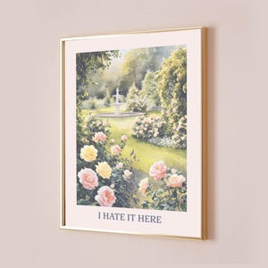 Puede incluir: Una impresión enmarcada de una pintura de acuarela que representa una escena de jardín con una fuente blanca, exuberante follaje verde y rosas rosadas y amarillas. El texto "I hate it here" está impreso debajo de la imagen.