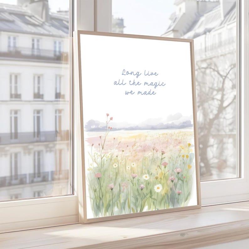 May include: A framed watercolor print featuring a field of wildflowers in soft pinks, yellows, and greens. The artwork includes the text "Long live all the magic we made" in a delicate blue script. The frame is a light wood color.