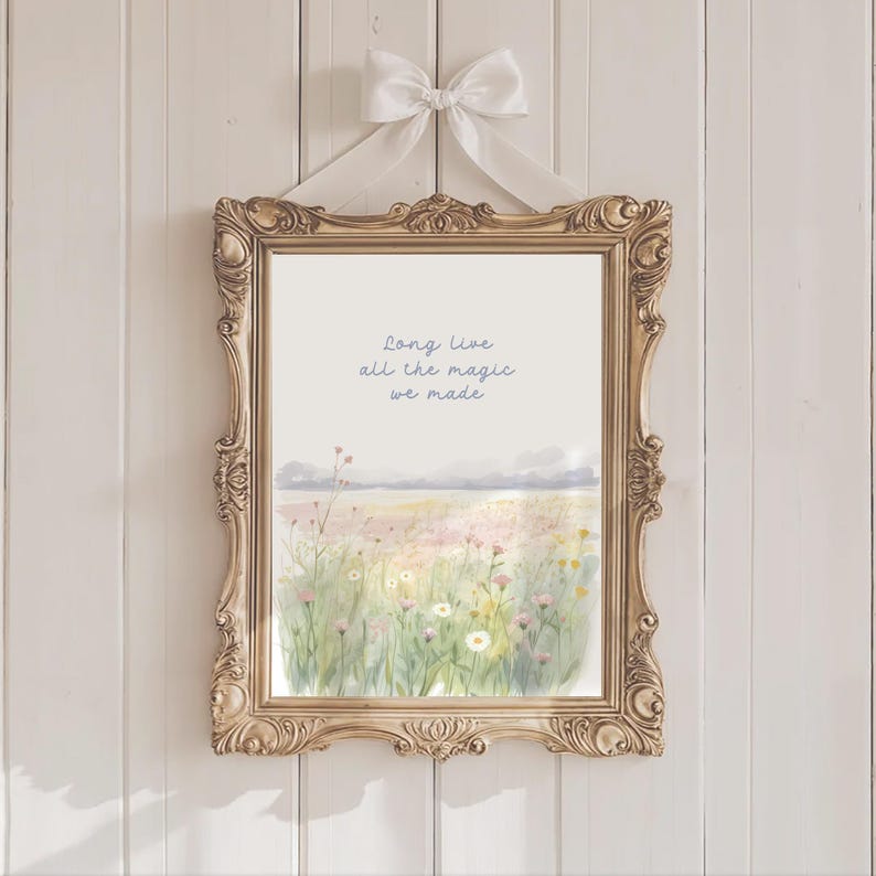 May include: A framed print with a watercolor landscape of a field of wildflowers, mountains, and the text "Long Live all the magic we made." The ornate gold frame is suspended by a white ribbon tied in a bow.