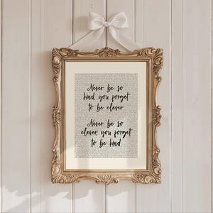 May include: A framed print with a gold ornate frame and a white ribbon. The print features the text "Never be so kind, you forget to be clever. Never be so clever, you forget to be kind" in black script over a background of smaller text.