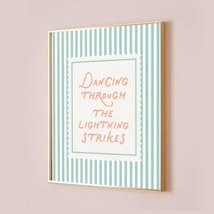May include: A framed art print with a gold-colored frame. The artwork features a light blue and white striped background with a white rectangle in the center. The text "DANCING THROUGH THE LIGHTNING STRIKES" is written in coral-colored script.