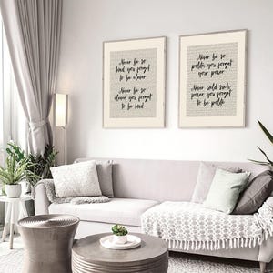 May include: Two framed art prints with inspirational quotes in black script on a cream background. The quotes are set against a backdrop of faint text. The frames are a light beige colour. The prints hang above a grey sofa.