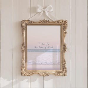 Peut inclure: Une impression encadrée avec une scène de plage et le texte "To live for the hope of it all." Le cadre doré orné est suspendu par un ruban blanc noué en nœud. L'impression présente une palette de couleurs pastel de rose, bleu et blanc.