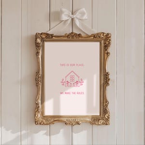 May include: A framed print with the text "THIS IS OUR PLACE. WE MAKE THE RULES." in pink. The print features a house illustration and floral accents. The ornate gold frame is suspended by a white ribbon and bow.