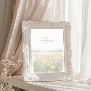 May include: A white framed art print featuring a watercolor landscape of a field of wildflowers. The print has the text "Long live all the magic we made" in light blue script. The frame has ornate detailing.