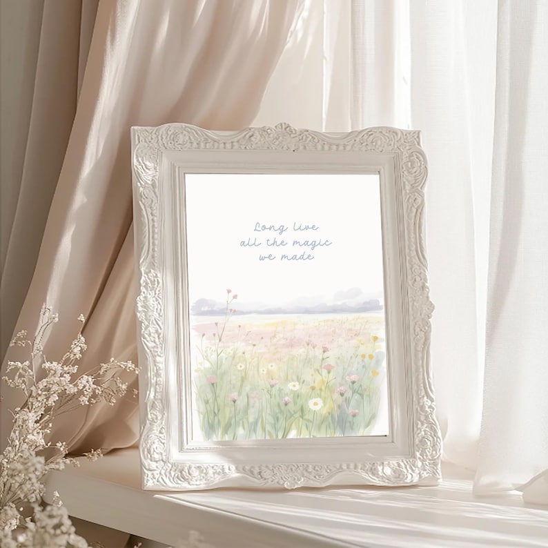 May include: A white framed art print featuring a watercolor landscape of a field of wildflowers. The print has the text "Long live all the magic we made" in light blue script. The frame has ornate detailing.