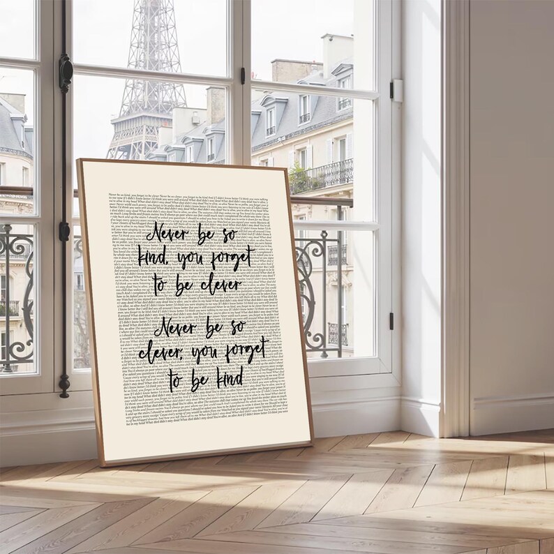 May include: A framed print featuring the text "Never be so kind, you forget to be clever. Never be so clever, you forget to be kind" in black script. The background is a collage of text. The frame is a light brown colour.