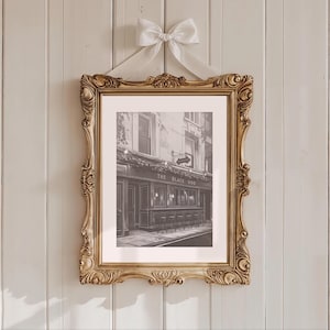 May include: A framed black and white print of a pub called "The Black Dog" with a gold ornate frame. The frame is suspended by a white ribbon tied in a bow. The pub has a dark facade with a sign.