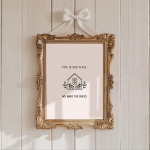 May include: A framed print with the text "THIS IS OUR PLACE. WE MAKE THE RULES." The print features a house illustration with floral accents. The ornate gold frame is suspended by a white ribbon tied in a bow.