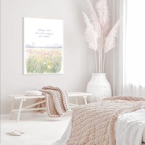 May include: A framed watercolor print with the text "long live all the magic we made" above a field of flowers. A white bench holds a beige knitted throw and a round pillow. A large white vase holds tall, feathery, pink plumes.
