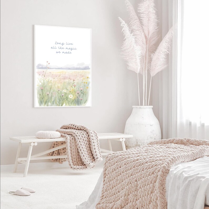 May include: A framed watercolor print with the text "long live all the magic we made" above a field of flowers. A white bench holds a beige knitted throw and a round pillow. A large white vase holds tall, feathery, pink plumes.