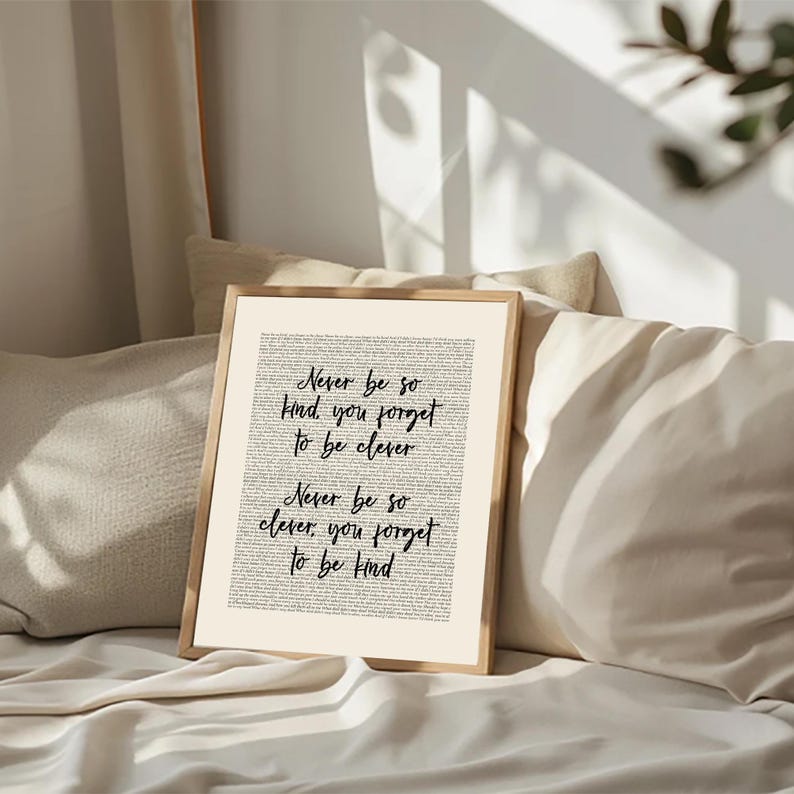 May include: A framed print featuring the quote "Never be so kind you forget to be clever. Never be so clever, you forget to be kind." The black script is on a cream background, within a light wood frame. The print is displayed on a bed with neutral-toned pillows.