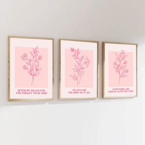 Op de afbeelding: Drie ingelijste kunstdrukken met roze botanische lijntekeningen op een lichtroze achtergrond. Elke print heeft een ander inspirerend citaat in een schreefletter. De frames zijn licht van kleur.