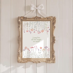 May include: A framed print with a gold ornate frame, hanging from a white ribbon bow. The print features watercolor flowers in pink, orange, and purple, with the text "Long live all the magic we made."