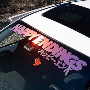 Peut inclure: Une voiture blanche avec un autocollant "HAPPY ENDINGS" rose et violet sur le pare-brise. L'autocollant comprend également le texte japonais "ハッピーエンド" en noir.