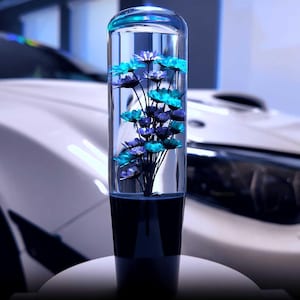 Peut inclure: Pommeau de levier de vitesse de voiture cylindrique transparent avec un bouquet de fleurs bleues et violettes à l'intérieur. La base est bleu foncé et le dessus est arrondi. Arrière-plan avec une voiture blanche.