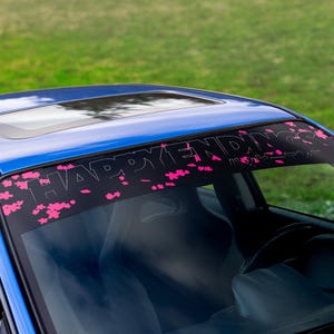 May include: A blue car with a black banner across the windscreen. The banner features the words "HAPPY ENDINGS" in white and pink cherry blossom designs. The car's interior is visible.