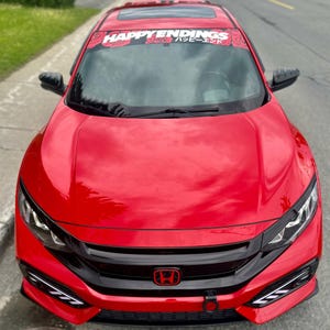 Op de afbeelding: Een felrode Honda Civic auto met zwarte accenten. Een banner over de voorruit leest "HAPPY ENDINGS" in het Engels en Japans. De auto heeft een rood Honda-embleem en een zwarte grille.