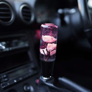 Könnte beinhalten: Ein transparenter Glas-Schalthebelknauf mit rosa Blumen, die in Harz eingebettet sind. Die Blumen sind durch das klare Glas sichtbar und von Glitzer umgeben.