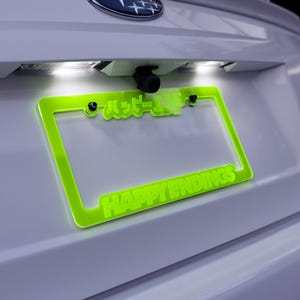 Könnte beinhalten: Ein grüner Nummernschildrahmen mit dem Text "HAPPY ENDINGS" und japanischen Schriftzeichen. Der Rahmen ist an einem weißen Auto befestigt.