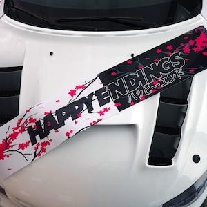 May include: A white car bonnet with a graphic overlay. The design features the words "HAPPY ENDINGS" in black and Japanese characters, alongside pink cherry blossom illustrations on a white and black background.