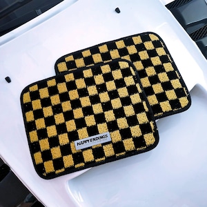 May include: Two black and gold checkered car floor mats. Each mat has a rectangular shape with rounded corners and a repeating pattern of black and gold squares. The mats have a label that reads "HAPPY ENDINGS".