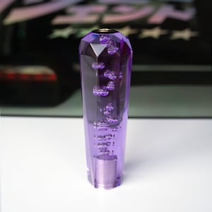 Op de afbeelding: Een paarse versnellingspookknop met een facetontwerp en interne bellen. De knop heeft een metalen basis en een goudkleurige bovenkant. De achtergrond is wazig, maar lijkt een voertuig te zijn.