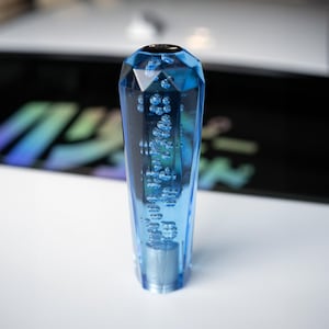 Puede incluir: Un pomo de cambio azul facetado con efecto burbuja. El pomo está hecho de material transparente azul claro con una base plateada. La parte superior del pomo es negra.