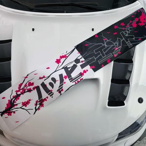 Könnte beinhalten: Eine weiße Motorhaube mit einem dekorativen Aufkleber mit japanischem Text und Kirschblütenzweigen. Der Aufkleber hat einen schwarz-weißen Hintergrund mit rosa Blüten und Text.