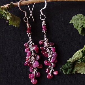 May include: A pair of dangle earrings with a silver chain and pink gemstone beads. The earrings are made with a silver hook and have a cluster of pink gemstones at the bottom.