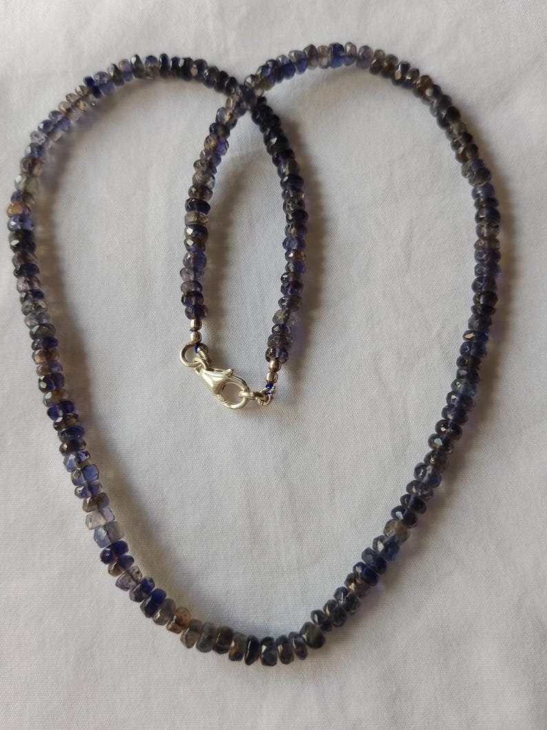 May include: A necklace made of small, faceted, dark blue gemstones. The necklace is strung on a silver chain and has a silver clasp.