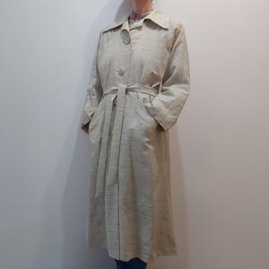 Può includere: Un trench coat beige chiaro con chiusura a bottoni, cintura e tasche. Il cappotto ha un collo e maniche lunghe. Il cappotto è lungo fino al ginocchio e sembra essere realizzato in un tessuto strutturato.