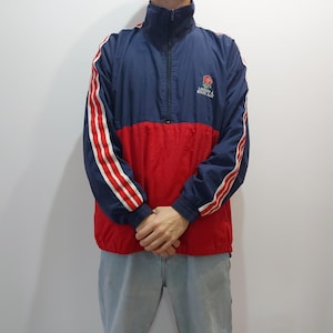 Op de afbeelding: Een blauw en rood windjack met witte strepen op de mouwen. Het jack heeft een ritssluiting en een opstaande kraag. Het jack heeft een logo op de linkerborst met de tekst "London Mounta".