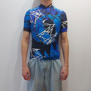 Op de afbeelding: Een blauw, zwart en wit fietsjersey met een geometrisch patroon. Het jersey heeft een rits en een kraag.