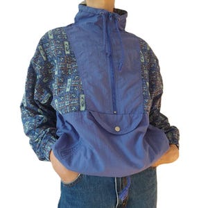 Puede incluir: Chaqueta anorak azul vintage con cuello alto y cierre de media cremallera. La chaqueta presenta un panel frontal azul liso y mangas estampadas con formas geométricas azules, marrones y turquesas. Tiene un bolsillo delantero y cierre de botón.
