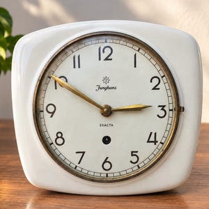 May include: A white ceramic table clock with a rounded square frame and a circular face. The clock features a brass-colored bezel, black Arabic numerals, and gold-toned hands. The dial displays the text Junghans and the word EXACTA.