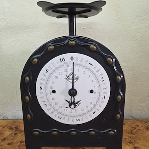 Op de afbeelding: Een zwarte mechanische keukenweegschaal met een witte ronde wijzerplaat met dubbele schaalverdeling voor kilogrammen en ponden. De weegschaal heeft een zwart metalen frame met messingkleurige klinknagels en een bovenste weegplateau.