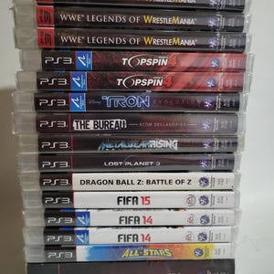 Könnte beinhalten: Ein Stapel PlayStation 3-Spiele. Zu den Titeln gehören "WWE Legends of WrestleMania", "Top Spin 4" und "FIFA 14". Die Hüllen sind transparent und zeigen Spieltitel und -grafiken. Verschiedene Sport- und Actionspiele.