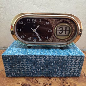 May include: A gold-toned, oval-shaped analog clock with a dark brown face and white hands. The clock displays the date as "31" and sits atop a blue box with a repeating diamond pattern. The clock's face has the word "DIAMOND".