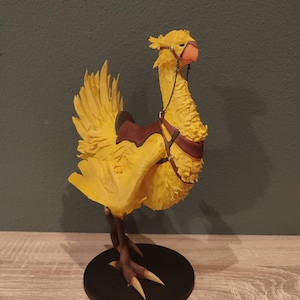 May include: A yellow 3D printed Chocobo figurine with a brown saddle and bridle. The Chocobo is standing on a black base with the text "BANANABREAD 3DCREATION" printed on a yellow base.