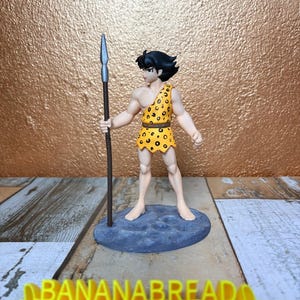 Può includere: Statuetta di un personaggio dei cartoni animati in un completo giallo con stampa leopardata, che tiene in mano una lancia. La figura è su una base blu. Il testo "BANANABREAD 3DCREATION" è in basso in giallo.