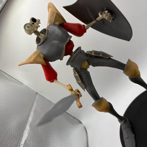 May include: A skeletal knight figurine with a sword and shield. The knight is wearing black and gold armor with a red tunic. The figurine is standing on a black base.